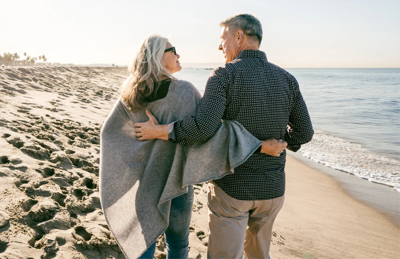 Imagen de dos personas jubiladas abrazadas paseando por la playa con una actitud de felicidad y complicidad