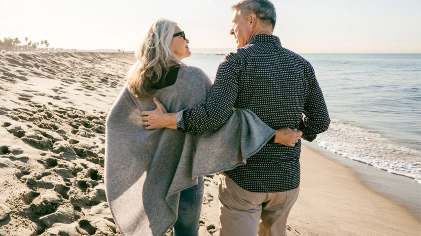 Imagen de dos personas jubiladas abrazadas paseando por la playa con una actitud de felicidad y complicidad